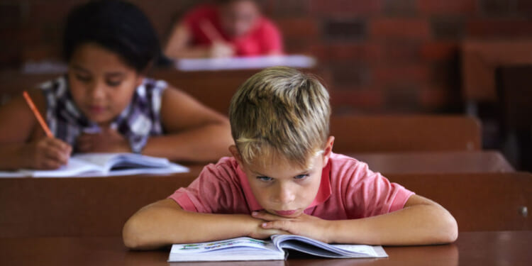 Bored, restless boy rests head on arms as he sits at his classroom desk, while female students behind him are working away.