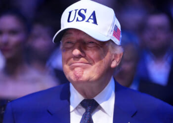 A grinning Donald Trump in a blue suit with white baseball cap with USA in blue lettering.