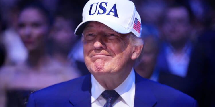 A grinning Donald Trump in a blue suit with white baseball cap with USA in blue lettering.