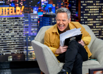 Greg Gutfeld sits in a gray armchair, laughing while holding papers and a clipboard. Behind him is a cityscape backdrop with the word GUTFELD! in bright lights.