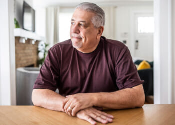 Middle-aged Hispanic man in a brown shirt sitting at a dining room table looking out toward a window.