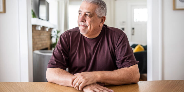 Middle-aged Hispanic man in a brown shirt sitting at a dining room table looking out toward a window.