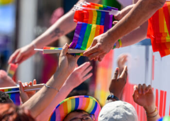 Someone leans over to hand a crowd of people rainbow pride flags during a parade.