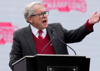Gov. Mike DeWine gestures from the podium with an NCAA backdrop behind him.