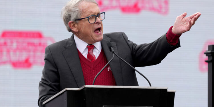 Gov. Mike DeWine gestures from the podium with an NCAA backdrop behind him.
