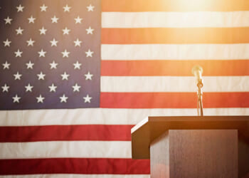A podium in front of an American flag. Bright light hangs above the microphone.