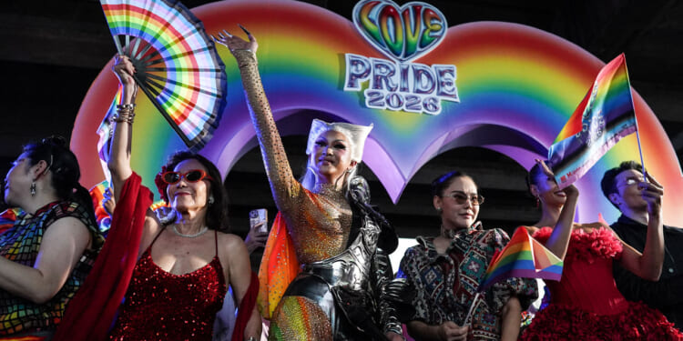 Colorfully-dressed individuals celebrate in front of a rainbow backdrop reading "Love PRIDE 2026)
