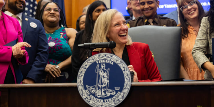 A laughing Abigail Spanberger sits at a desk surrounded by smiling, laughing people.
