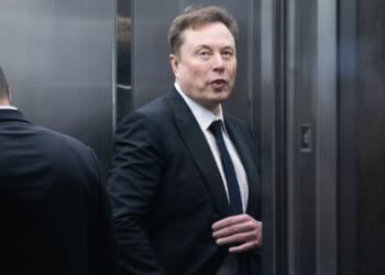 Elon Musk in a dark suit looks toward camera.