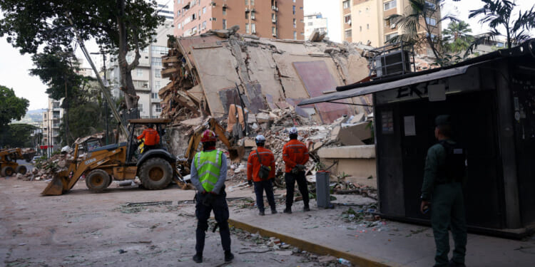 Several workers stand in front of the rubble of a collapsed building as a bulldozer works with the rubble.