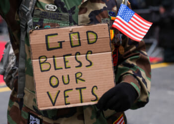Individual in military fatigues holds a small flag and a cardboard sign reading "God Bless Our Vets."