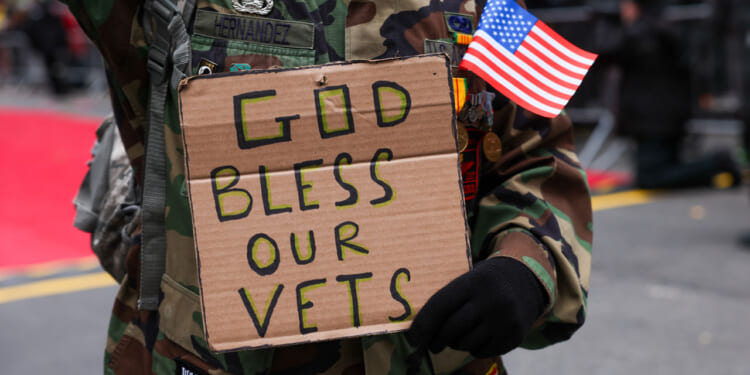 Individual in military fatigues holds a small flag and a cardboard sign reading "God Bless Our Vets."