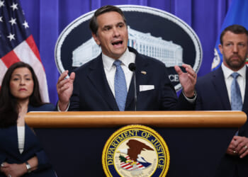 A man in a suit speaks at a podium with the Department of Justice seal, flanked by a woman and another man, both in business attire. American flags and an official emblem are visible in the background.