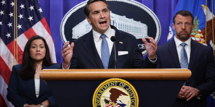 A man in a suit speaks at a podium with the Department of Justice seal, flanked by a woman and another man, both in business attire. American flags and an official emblem are visible in the background.