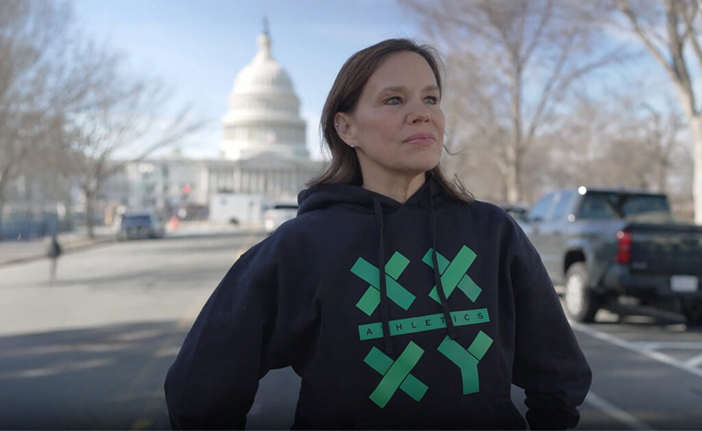 A woman in a black hoodie with green text stands on a street with the U.S. Capitol building visible in the background on a clear day.
