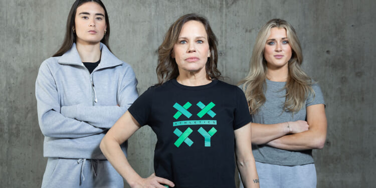 Three women stand in front of a concrete wall. The woman in the center, Jennifer Sey, wears a black XX-XY Athletics t-shirt, while the other two, Paula Scanlan and Riley Gaines in gray outfits, stand on either side with arms crossed. All face the camera with serious expressions.