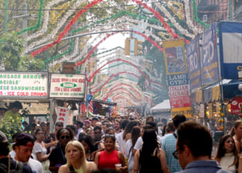 A crowded Italian street festival with food stalls, colorful hanging decorations, and many people walking and enjoying the event on a sunny day.