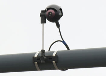 A black surveillance camera is mounted on a metal pole with brackets and a cable attached, set against a plain, light-colored background.