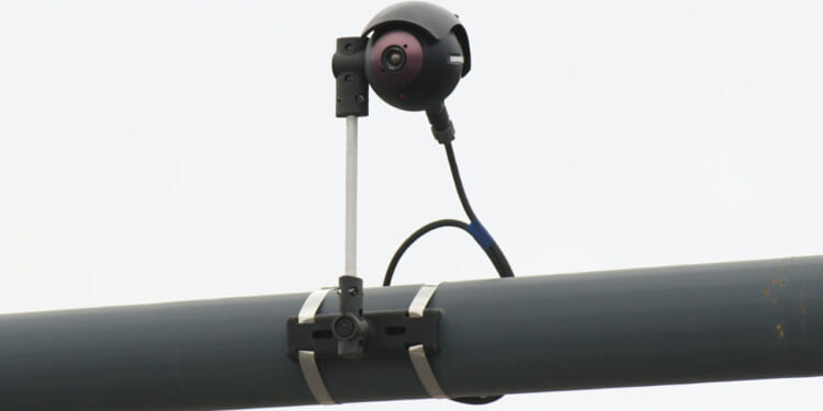 A black surveillance camera is mounted on a metal pole with brackets and a cable attached, set against a plain, light-colored background.