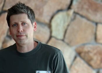 A man with short brown hair wearing a dark green shirt stands in front of a wall made of large, irregular stone blocks.