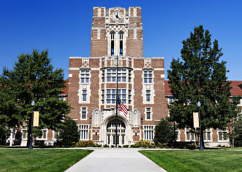 The University of Tennessee, Knoxville: a large brick university building with a central clock tower, arched entrance, and an American flag in front, surrounded by green trees and a clear blue sky.