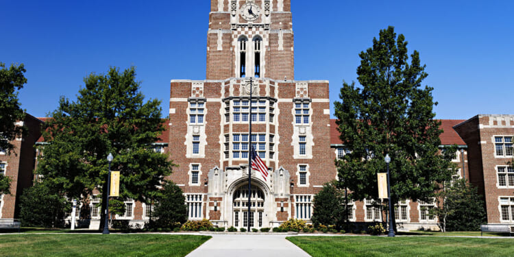 The University of Tennessee, Knoxville: a large brick university building with a central clock tower, arched entrance, and an American flag in front, surrounded by green trees and a clear blue sky.
