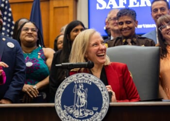 A smiling woman in a red blazer sits at a desk with the Virginia state seal, surrounded by happy people, at what appears to be a government event, with a “SAFE” sign partially visible behind them.