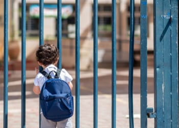 A child with a school backpack look on schoolyard grounds toward an open entrance door.