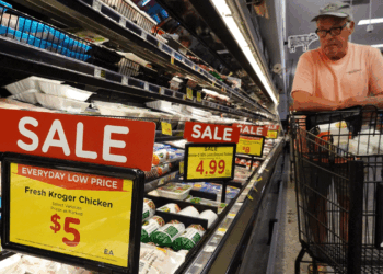 A man in glasses and a hat stands by a grocery cart in a store aisle, looking at sale signs for fresh Kroger chicken.