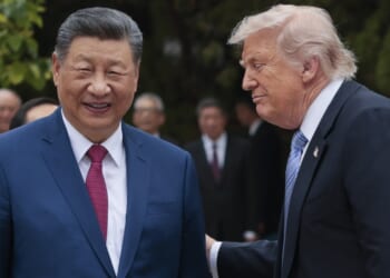Two men in suits stand outdoors among a group of people, one smiling and the other placing a hand on his back. Trees and greenery are visible in the background.