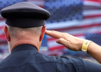 A police officer with a security wristband salutes in front of a large, blurred American flag backdrop, proudly honoring America.