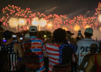 People dressed in American flag-themed clothing watch colorful fireworks light up the night sky, celebrating together outdoors.