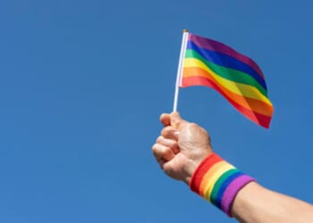 A person holds a LGBTQ+ flag up in the air while wearing a wristband showcasing the same colors.