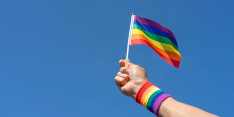 A person holds a LGBTQ+ flag up in the air while wearing a wristband showcasing the same colors.