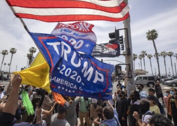 A crowd gathers outdoors by a street lined with palm trees, waving flags including a large Trump 2020 banner.