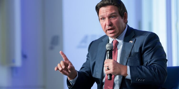 A man in a blue suit and red tie speaks into a microphone while gesturing with his hand during an indoor event.
