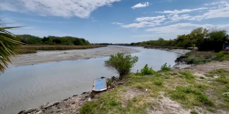 A river with low water levels winds through a grassy landscape under a partly cloudy sky. Vegetation lines the banks, and a small wooden structure sits near the water’s edge in the foreground.