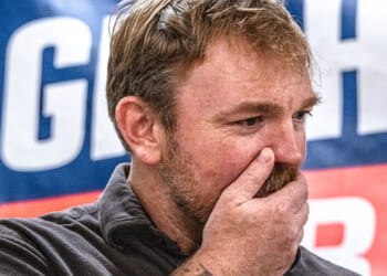 A man with light brown hair and a beard covers his mouth with his hand, appearing thoughtful or concerned. A blue banner with large white letters is blurred in the background.