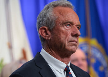 A man in a suit stands in front of blue and white flags, looking serious. The background is blurred, and he is facing left with a focused expression.