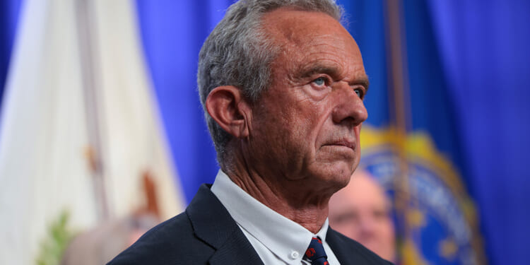 A man in a suit stands in front of blue and white flags, looking serious. The background is blurred, and he is facing left with a focused expression.