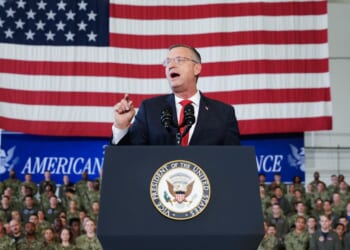 A man in a suit speaks at a podium with the U.S. Vice Presidential seal. An American flag and a group of uniformed military personnel are visible in the background.