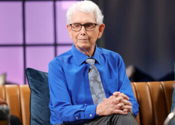 Walt Heyer in a blue shirt and light blue tie sits with his hands folded on the set of a TV show.