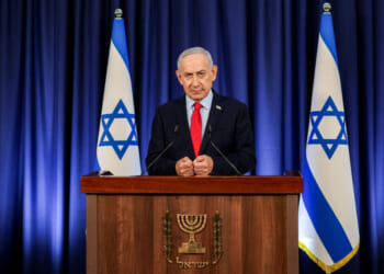 Benjamin Netanyahu stands at a podium with the Israeli emblem, flanked by two Israeli flags, delivering a speech about Israel in front of a blue curtain.