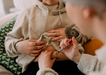 A person injects IVF medication into their abdomen while another supports them. Both have tattoos. They are sitting on a couch with a patterned pillow.