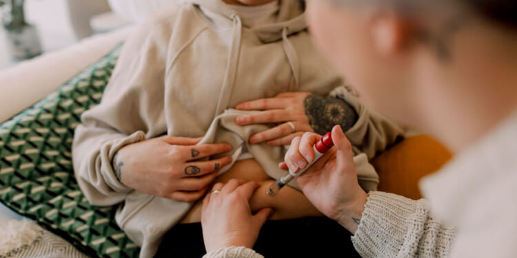 A person injects IVF medication into their abdomen while another supports them. Both have tattoos. They are sitting on a couch with a patterned pillow.