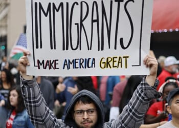 A person in a crowd holds a sign reading Immigrants make America great. People are gathered in the background, some holding flags, at an outdoor event or protest.