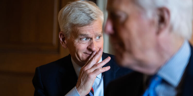 A man with short gray hair in a suit smiles with his hand near his mouth, while another man in a suit is blurred in the foreground. They appear to be having a conversation indoors.