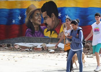 Four people walk past rubble in front of a mural featuring two people, a woman in a hat and a man in yellow, with a Venezuelan flag backdrop.