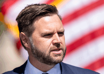 JD Vance, a man with short brown hair and a beard, wearing a suit and tie, stands outdoors in front of a blurred American flag.
