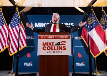 A man stands at a podium labeled Mike Collins U.S. Senate, arms outstretched, with American and Georgia state flags behind him and campaign signs in the background.